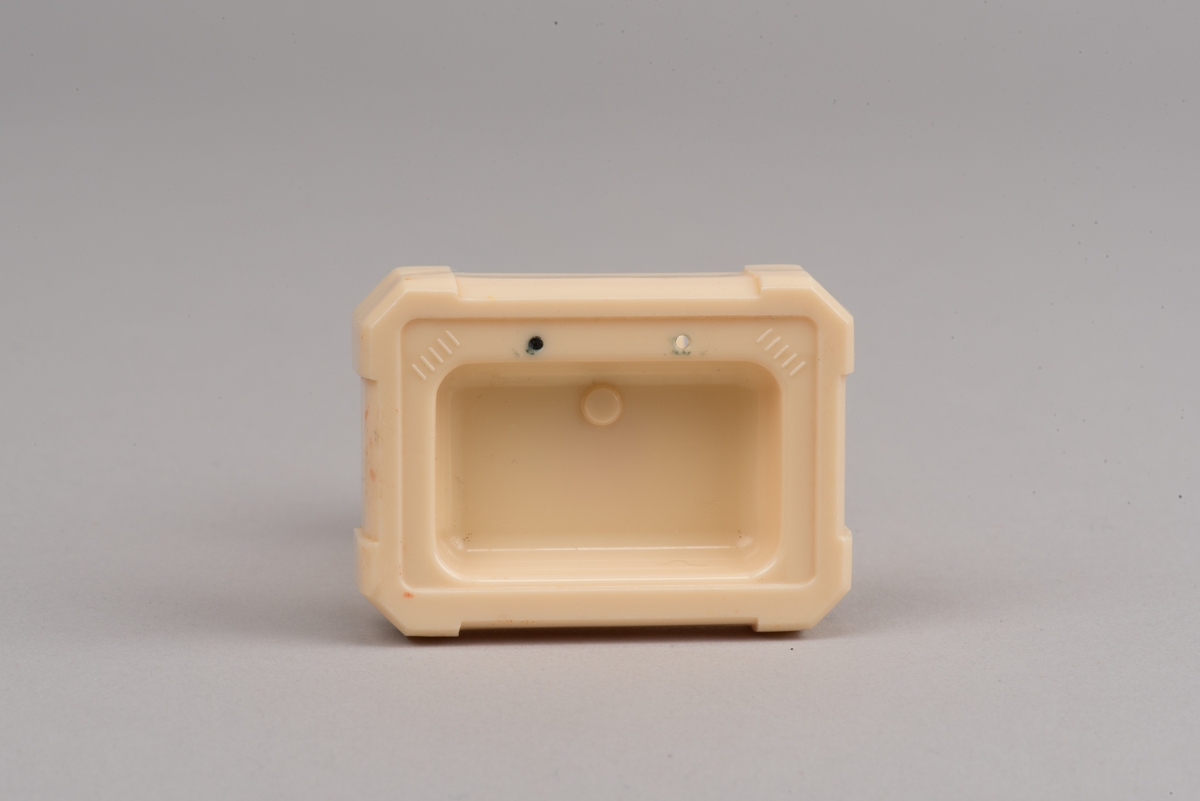Dockskåpsinredning i form av ett tvättställ av beige plast. 
Fyrkantigt handfat med avfasade hörn på avsmalnande pelare. Kranarna saknas men det finns två hål för dessa. vid sidorna av kranarna finns två räfflade partier i svag relief, för tvålar. Skala 1:18
