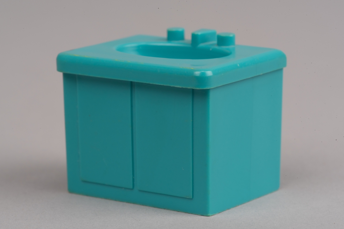Dockskåpinredning i form av ett turkost tvättställ av plast.
Tvättstället är rektangulärt och har två dörrar på framsidan i relief. Handfatet sticker ut lite på framsidan och har en oval försänkning. Bakom denna två kranar och en blandare i förenklad form. I botten ett hål för vattenavrinning.