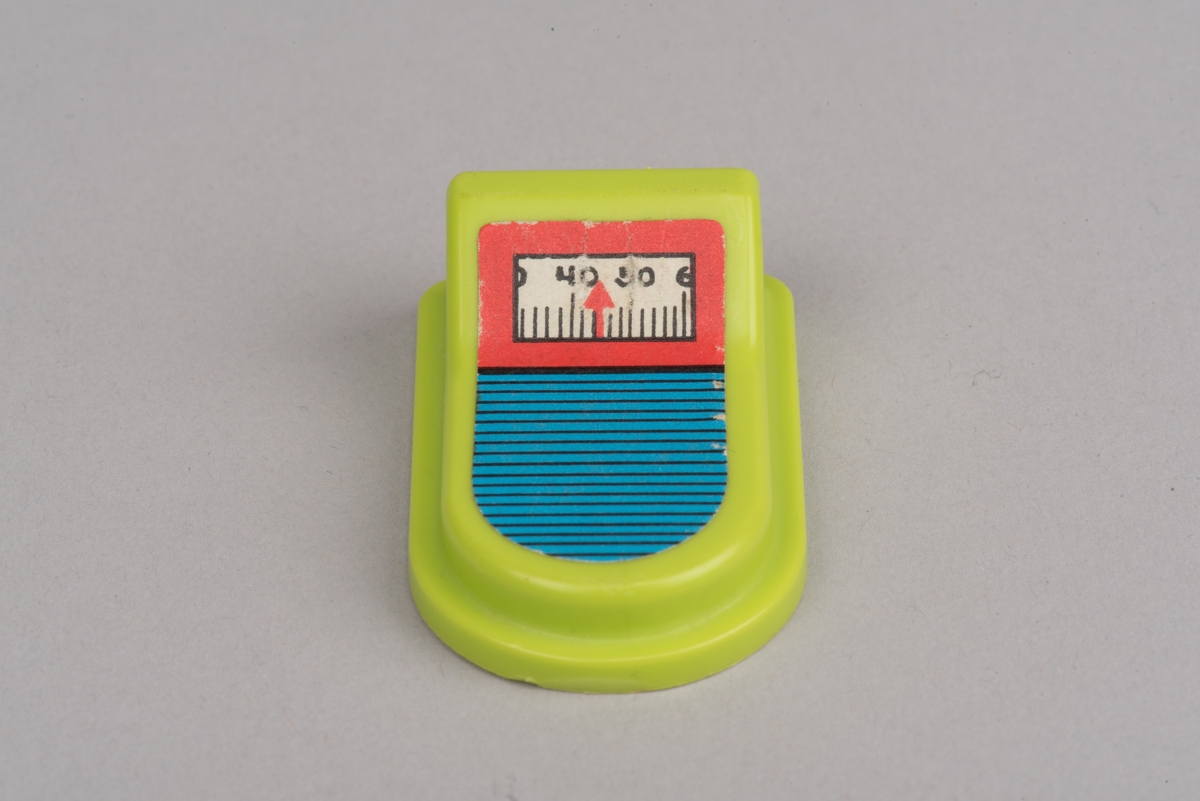 Dockskåpsinredning i form av en personvåg av gulgrön plast.
Rektangulär våg med rundad framkant och förhöjt bakstycke. På ovansidan sitter en fastlimmad etikett med en skala från 30 till 60 samt gradstreck. Själva "vågen" är blå med svarta ränder.