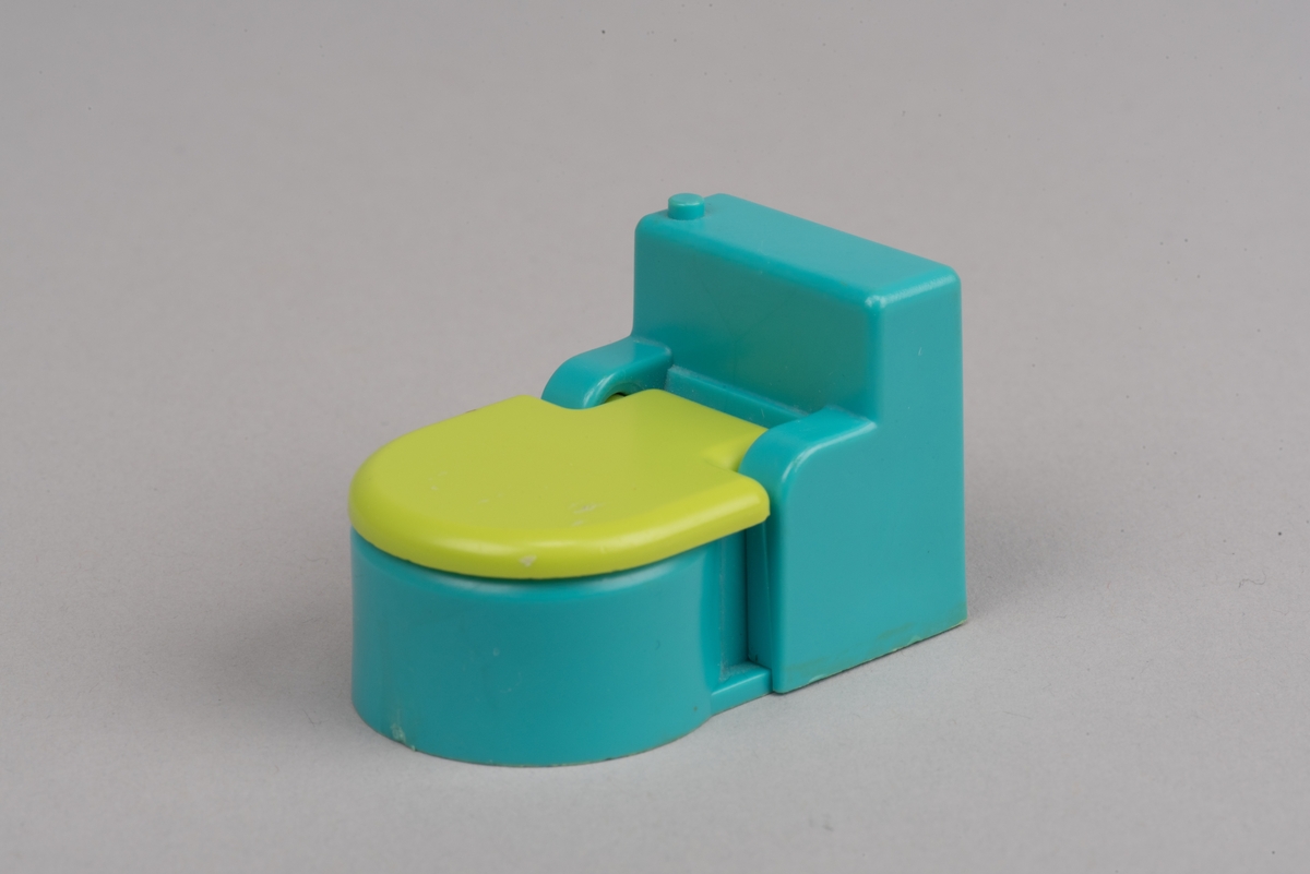 Dockskåpsinredning i form av en toalettstol av plast. Toalettlocket går att fälla upp.
Vattenbehållaren och stolen är turkosa. På ovansidan av behållaren finns en förhöjd spolknapp. Toalettlocket är gulgrönt.