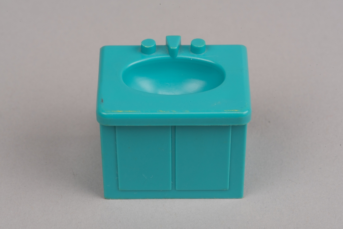 Dockskåpinredning i form av ett turkost tvättställ av plast.
Tvättstället är rektangulärt och har två dörrar på framsidan i relief. Handfatet sticker ut lite på framsidan och har en oval försänkning. Bakom denna två kranar och en blandare i förenklad form. I botten ett hål för vattenavrinning.