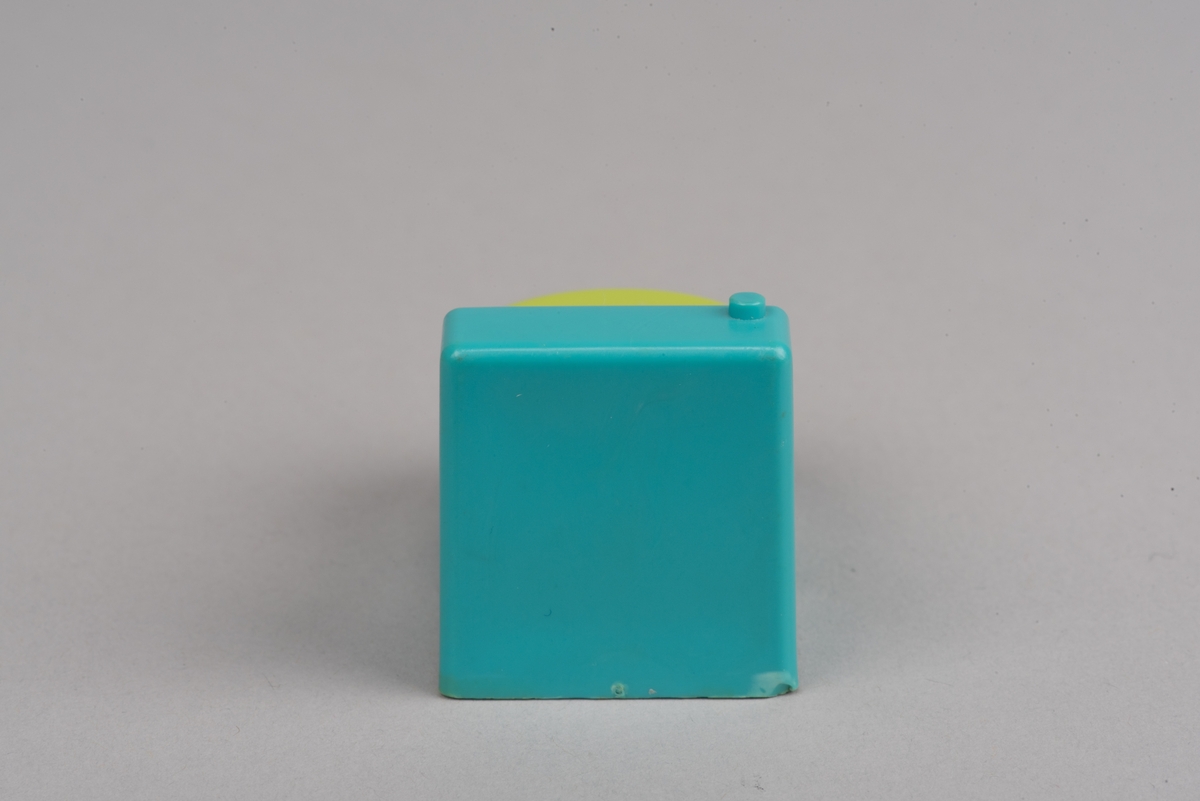 Dockskåpsinredning i form av en toalettstol av plast. Toalettlocket går att fälla upp.
Vattenbehållaren och stolen är turkosa. På ovansidan av behållaren finns en förhöjd spolknapp. Toalettlocket är gulgrönt.