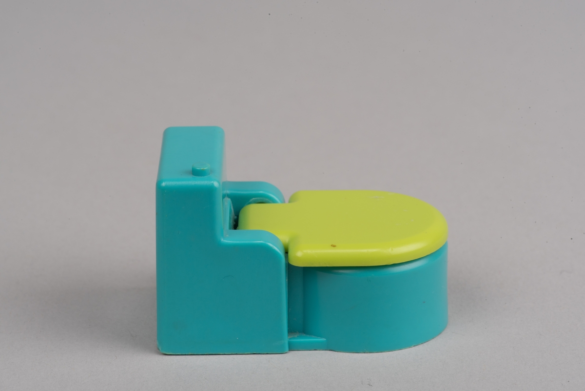 Dockskåpsinredning i form av en toalettstol av plast. Toalettlocket går att fälla upp.
Vattenbehållaren och stolen är turkosa. På ovansidan av behållaren finns en förhöjd spolknapp. Toalettlocket är gulgrönt.