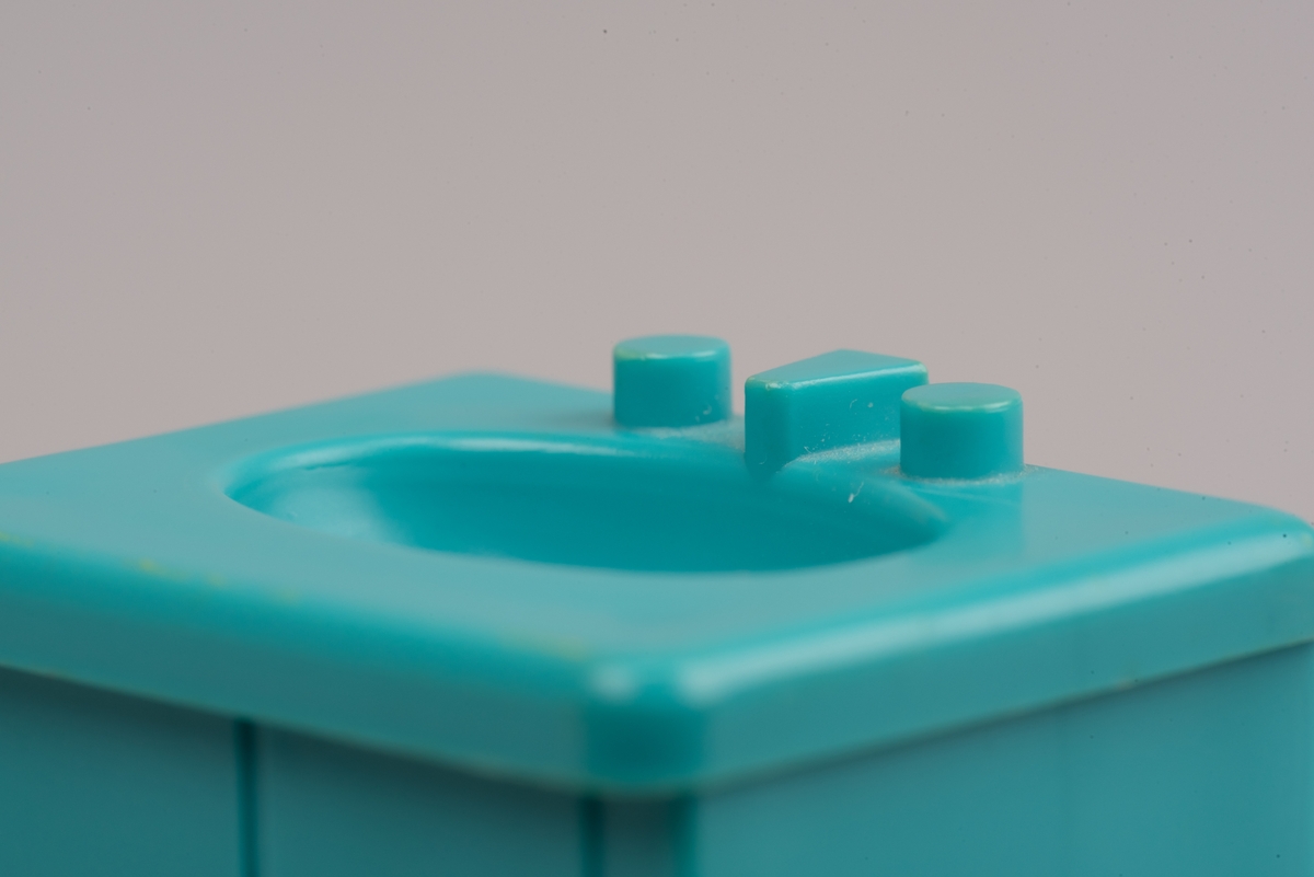 Dockskåpinredning i form av ett turkost tvättställ av plast.
Tvättstället är rektangulärt och har två dörrar på framsidan i relief. Handfatet sticker ut lite på framsidan och har en oval försänkning. Bakom denna två kranar och en blandare i förenklad form. I botten ett hål för vattenavrinning.