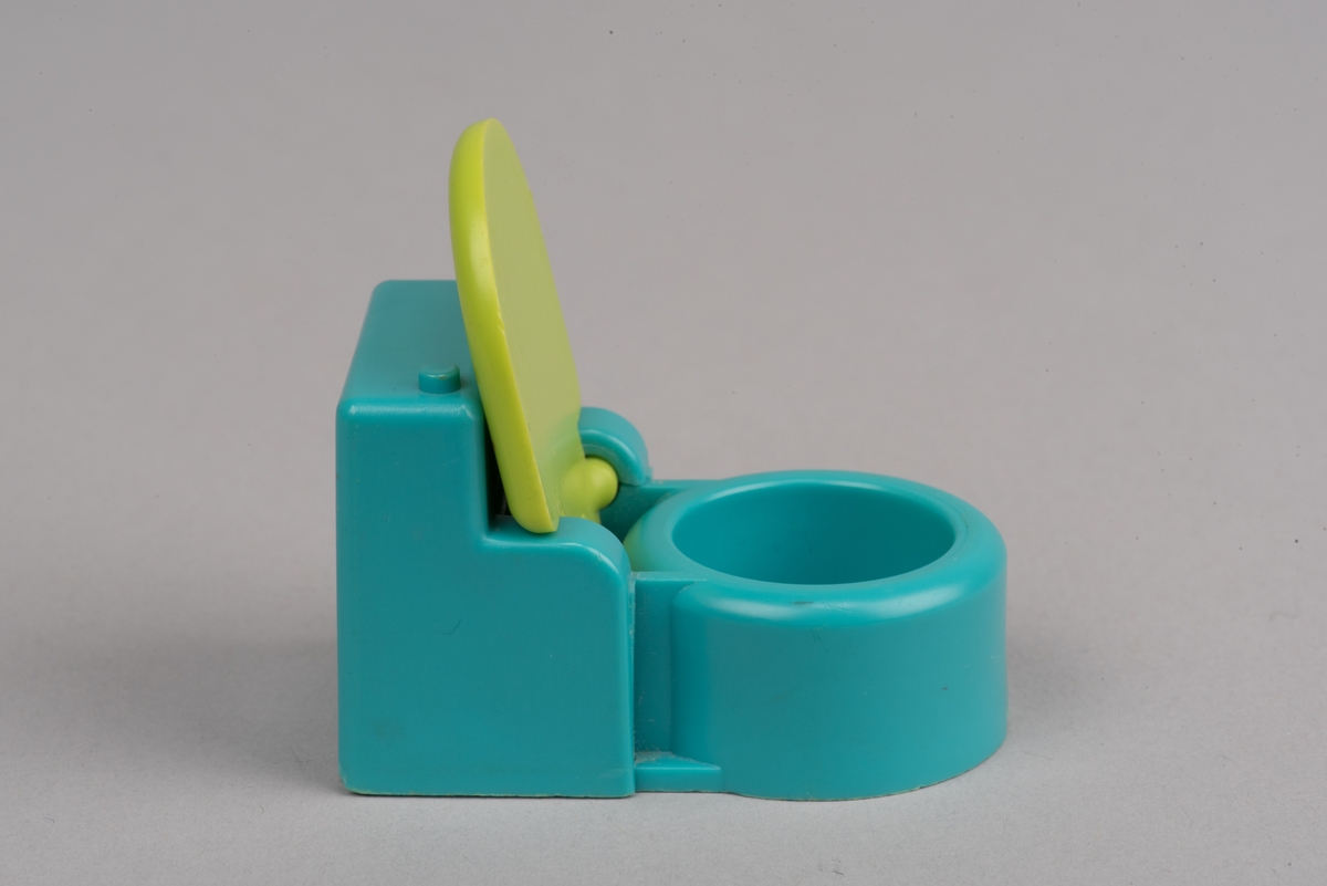 Dockskåpsinredning i form av en toalettstol av plast. Toalettlocket går att fälla upp.
Vattenbehållaren och stolen är turkosa. På ovansidan av behållaren finns en förhöjd spolknapp. Toalettlocket är gulgrönt.