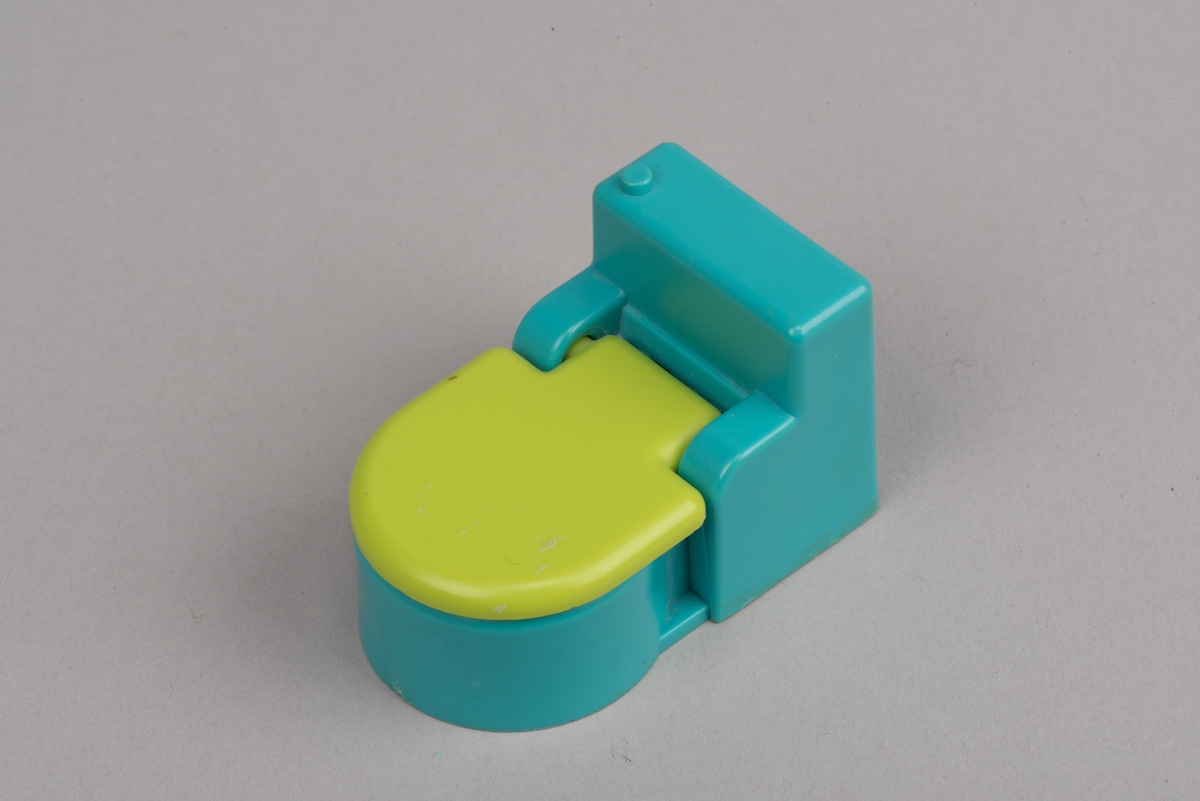 Dockskåpsinredning i form av en toalettstol av plast. Toalettlocket går att fälla upp.
Vattenbehållaren och stolen är turkosa. På ovansidan av behållaren finns en förhöjd spolknapp. Toalettlocket är gulgrönt.