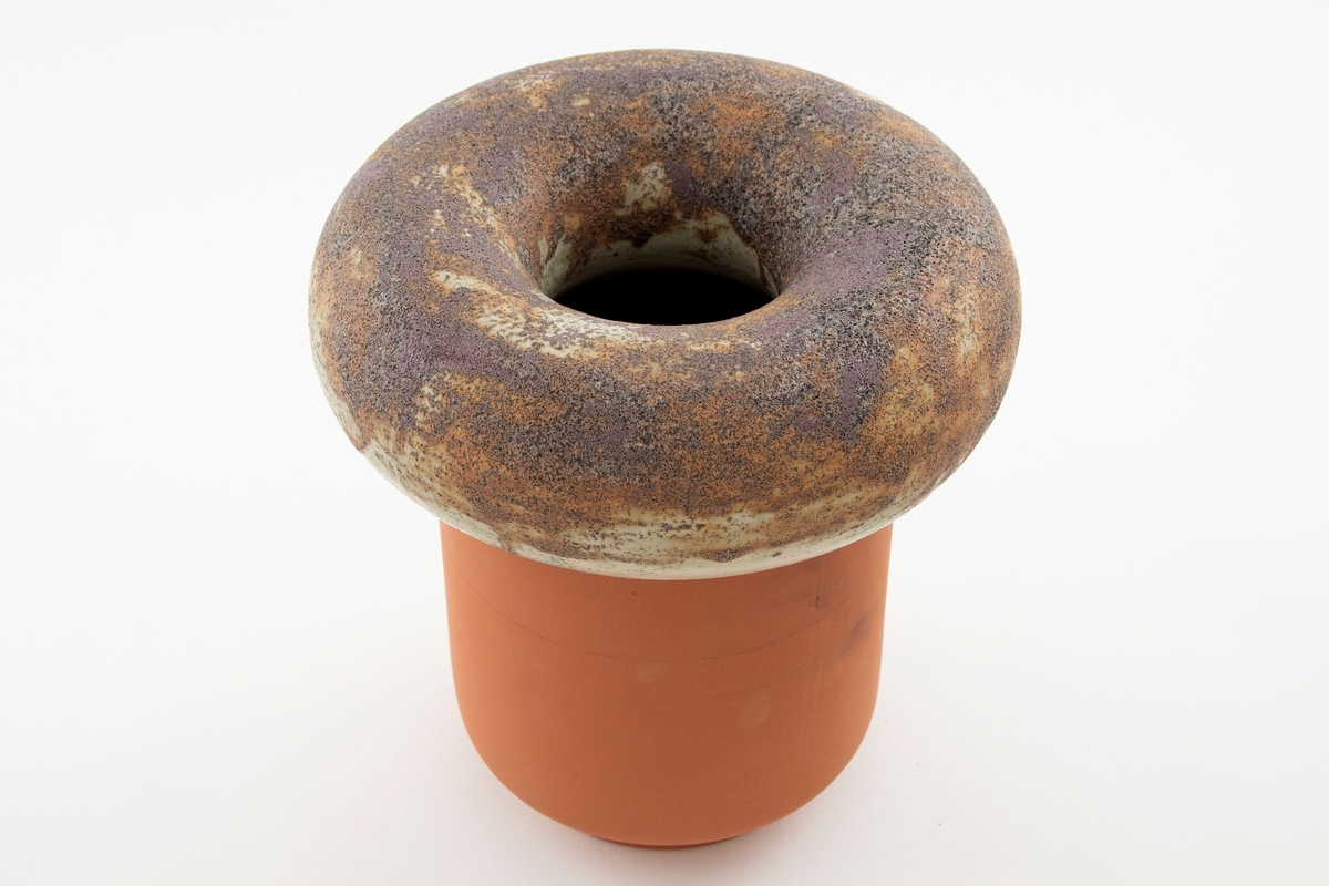 Vase i matt brun og beige terrakotta. Formen er satt sammen av en sylinder med lav fot og en voluminøs ring som ligger på toppen. Åpning i midten av ringen. Ringen er glasert i ulike matte brun-nyanser med grov tekstur.