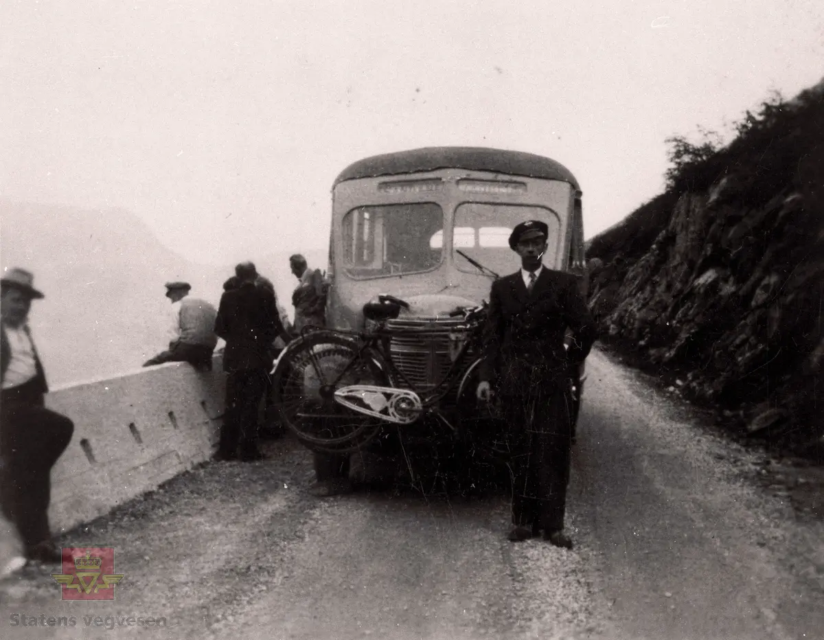 Ein rutebuss frå Firda Billag på vegen mellom Sandane og Vadheim. Frampå vogna heng to syklar. 
Lastebilen er en Volvo "rundnese" 1946-53. (Opplysninger til kjøretøyet fra Ivar E. Stav).

Rutevogna og passajerane har pause og støypekanten på staden var høg.