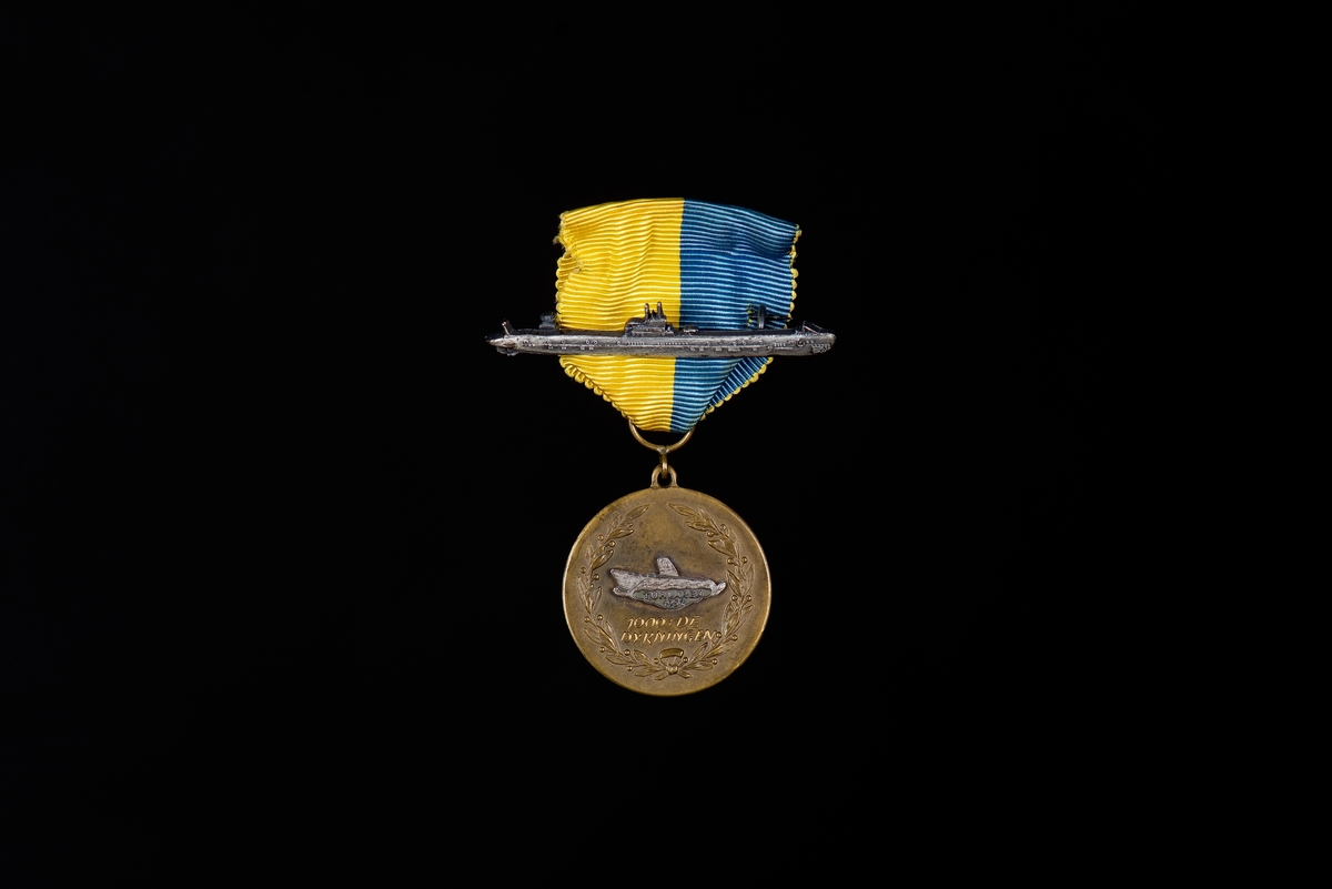 Medalj av brons. Rund med motiv av lagerkrans samt text: 1000:e dykningen" emblem i vitmetall i form av ubåten Tumlaren ingraverat med texten: "Tumlaren 1952". Medaljen baksida har text ingraverad: "SKNR2 FURIR ÖSTERHOLM". Medaljen hänger i blå-gult sidenripsband med säkerhetsnål fastsydd. På sidenbandet är en nål i form av ubåt fäst. Text på baksida av nålen: "SPORRONG & CO". Ubåten som avbildas är ubåt av Bävern-klass sjösatta 1921.