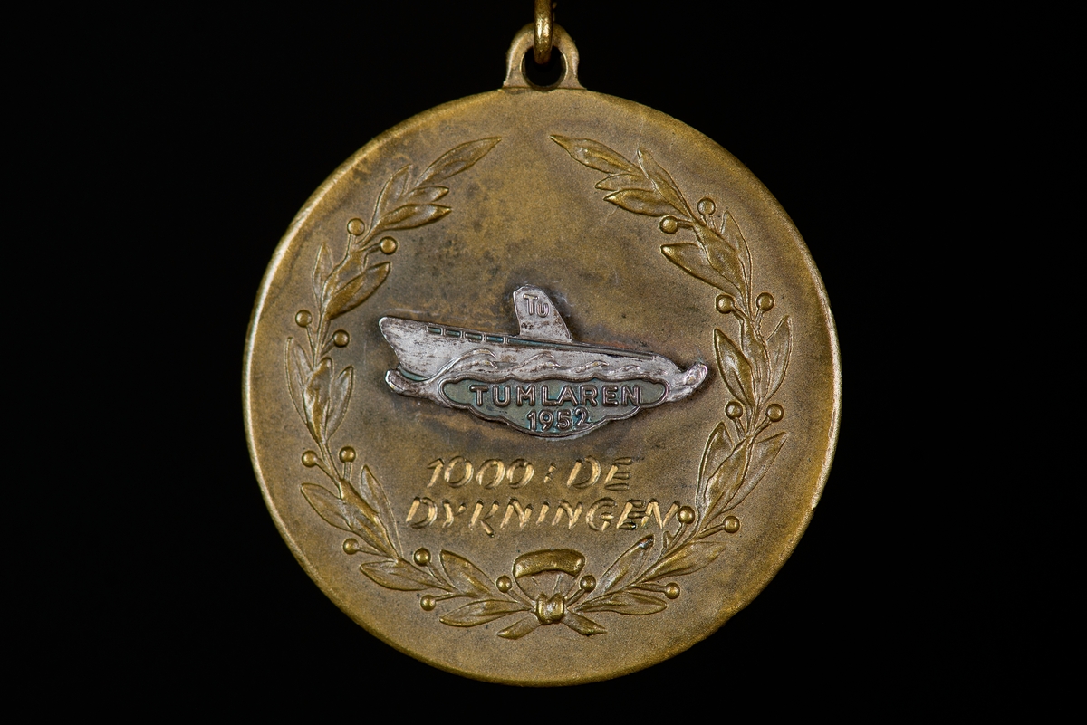 Medalj av brons. Rund med motiv av lagerkrans samt text: 1000:e dykningen" emblem i vitmetall i form av ubåten Tumlaren ingraverat med texten: "Tumlaren 1952". Medaljen baksida har text ingraverad: "SKNR2 FURIR ÖSTERHOLM". Medaljen hänger i blå-gult sidenripsband med säkerhetsnål fastsydd. På sidenbandet är en nål i form av ubåt fäst. Text på baksida av nålen: "SPORRONG & CO". Ubåten som avbildas är ubåt av Bävern-klass sjösatta 1921.