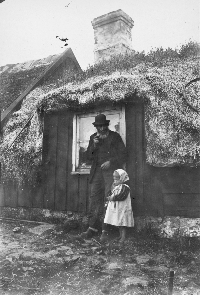 En piprökande man och en liten flicka står framför ett ovanligt stort fönster till en ryggåsstuga. Det är "Bolsestugan" i Trönninge som Severin Nilson 1908 köpte till målarateljé. Notera den ovanliga taktäckningen av ålgräs som vitnar när det torkat och kräver tjocka lager för att hålla tätt. Stugan är en högloftsstuga med tillbyggda loft vid båda gavlarna.
(Se även bildnr VMA6719_27.)