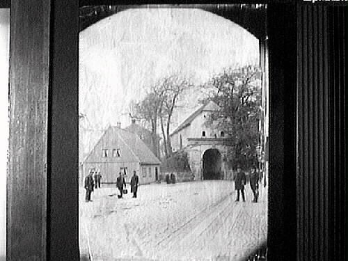 Gatubild vid Norre Port i Halmstad, sedd från Storgatan. Flera män i olika munderingar står längs vägen, vända mot fotografen. Den norra stadsporten ingick i det befästa Halmstad och stod klar 1601, alltså under danskt styre. Dess sneda vinkel gentemot Storgatan lär ha funktionen att förhindra beskjutning in mot staden. Varje natt stängdes alla stadsportarna till skydd åt invånarna. 

Stadsfullmäktige beslöt 1879 att riva portbyggnaden, men dåvarande riksantikvarien sa nej. Istället inrättade Hallands Museiförening (bildad 1886) där ett museum som öppnades 1899 och stängdes 1933. Norre Port blev byggnadsminne 2007.