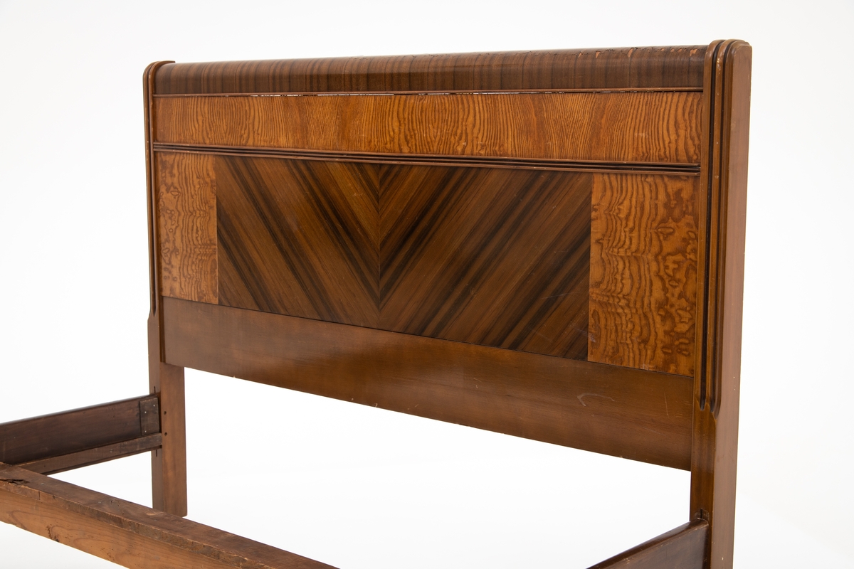 Seng i Art Deco stil. Består av hodegavl, endegavl, to sidestykker og tre tverrstykker til sengebunn. Hodegavlen er høyere enn endegavlen. Gavlstykkene er dekorert med felt i ulike typer finér, antatt bjørk og mahogni. Gavlene har avrundet kant på toppen i mørk finér, et felt med mørk finér i senter med finéren lagt i fiskebensmønster, som er rammet inn av et felt med lysere finér på hver side. Benene har påmontert små hjul. Skadet finér på hodegavl.