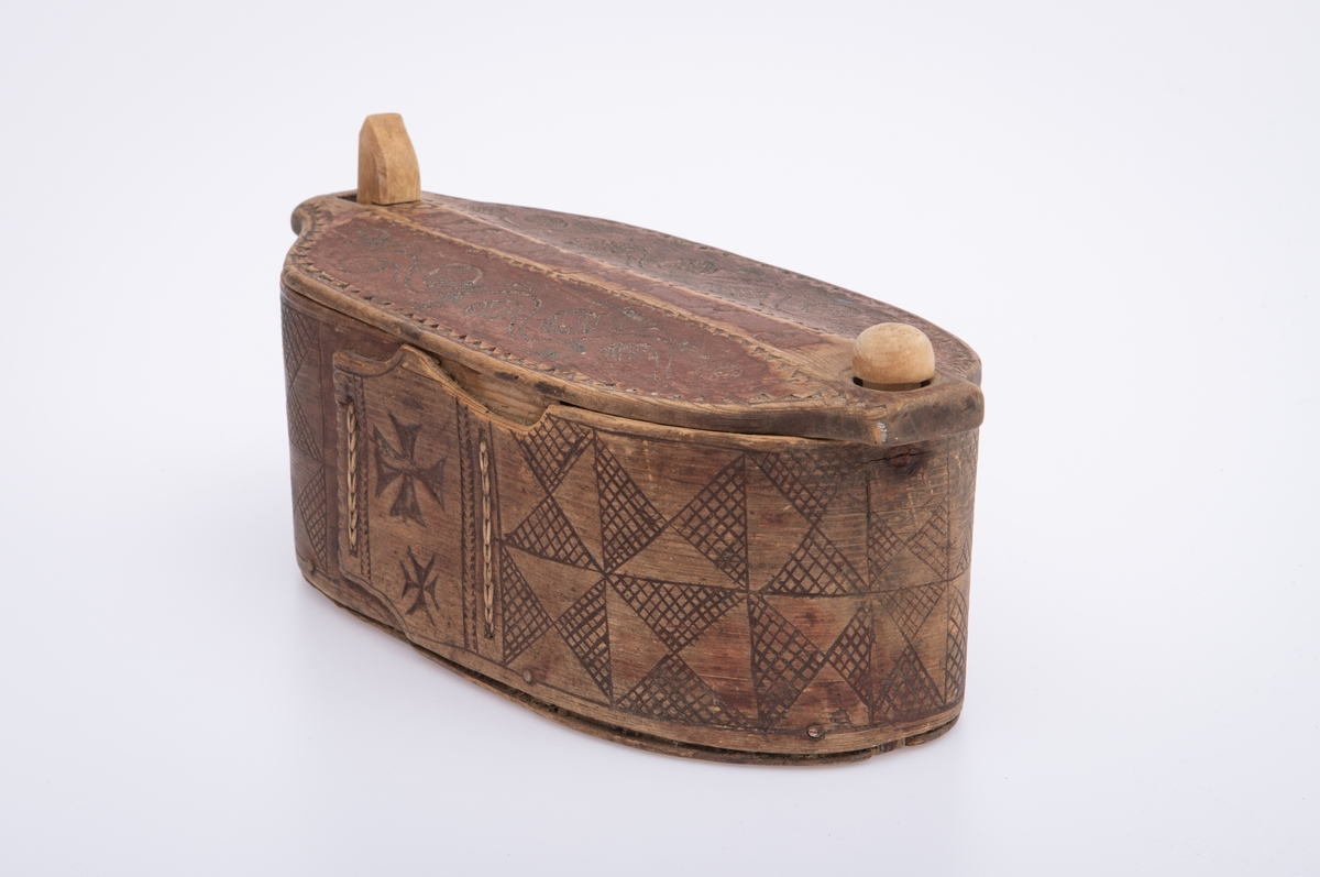 Furu, Furuspon bøyd kring oval botn og sydd manneh
med tæger. Skåret dekor, ranker og ruter. 2 jernkors mellom saumene i sida. Rep. fester for lokket. Rødmalt.