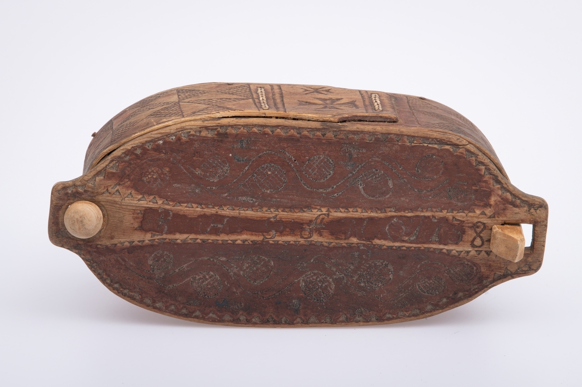 Furu, Furuspon bøyd kring oval botn og sydd manneh
med tæger. Skåret dekor, ranker og ruter. 2 jernkors mellom saumene i sida. Rep. fester for lokket. Rødmalt.