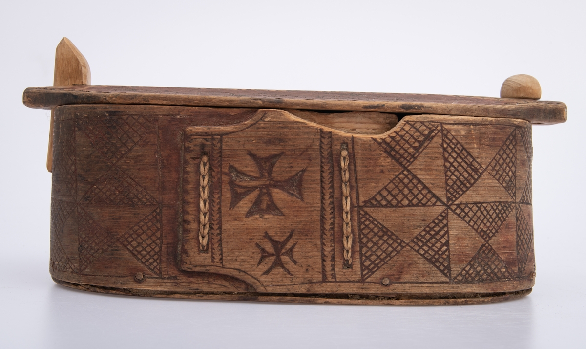 Furu, Furuspon bøyd kring oval botn og sydd manneh
med tæger. Skåret dekor, ranker og ruter. 2 jernkors mellom saumene i sida. Rep. fester for lokket. Rødmalt.