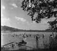 Barn och vuxna badar i en sjö. På strandkanten ligger en motorbåt med åror.