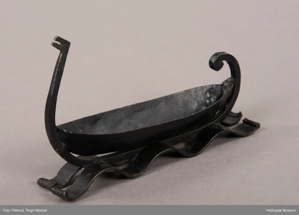 Et askebeger i form av et vikingskip plassert på to remser i jern i form av bølger. Laget av sort jern. Et enkelt utformet dragehode i baugen, i ene enden av et jernstykke som er montert på under båtformen og ender i en fremoverbøyd spiral i enden.