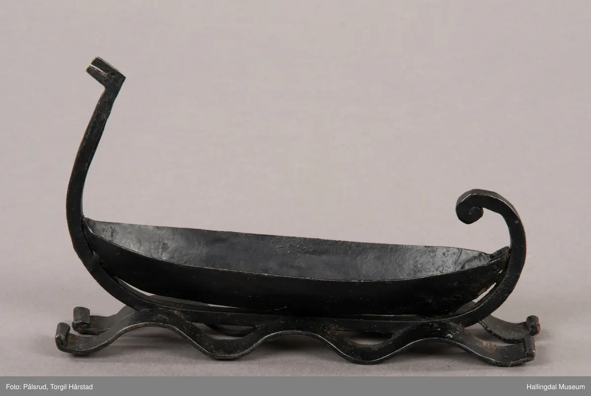 Et askebeger i form av et vikingskip plassert på to remser i jern i form av bølger. Laget av sort jern. Et enkelt utformet dragehode i baugen, i ene enden av et jernstykke som er montert på under båtformen og ender i en fremoverbøyd spiral i enden.