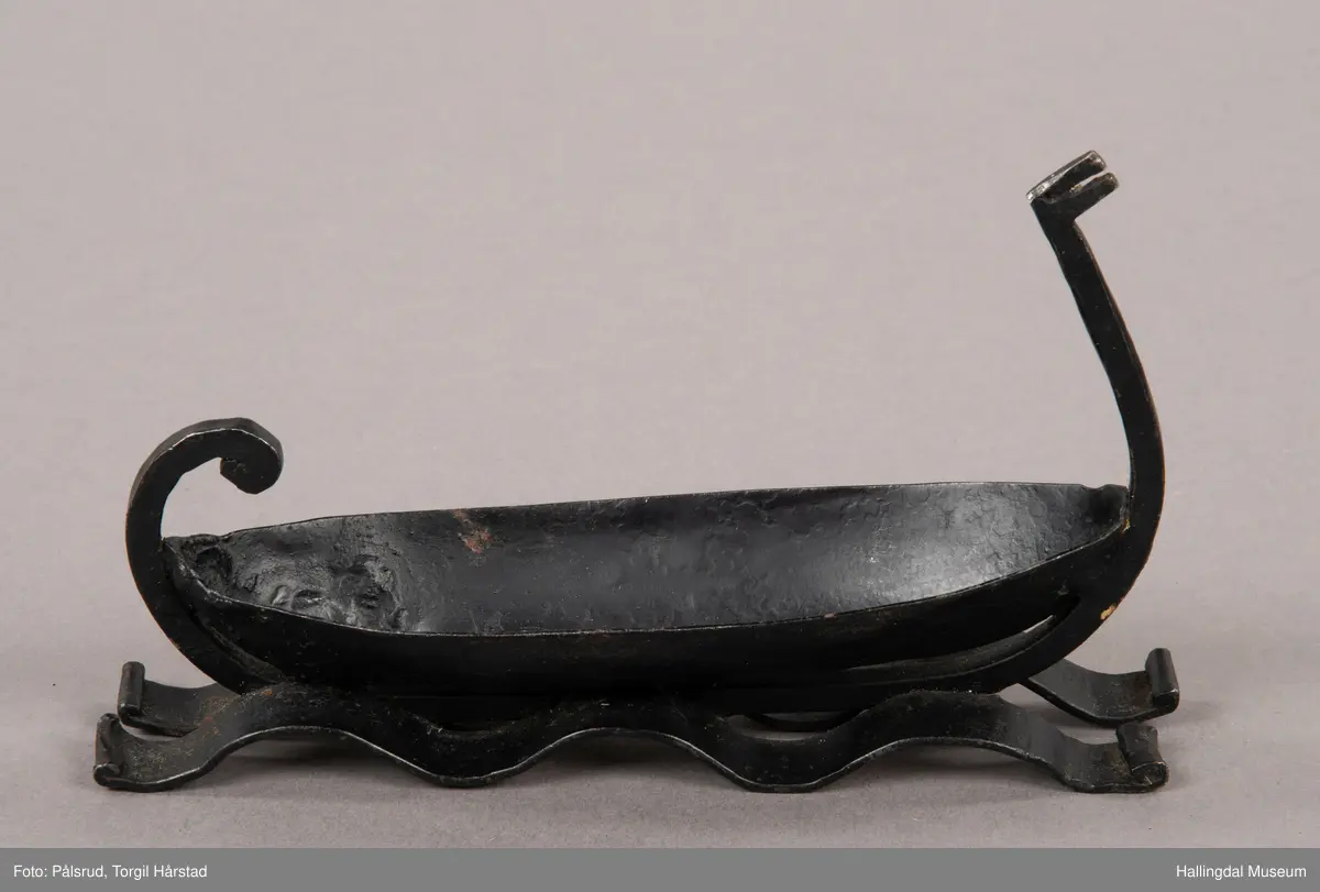 Et askebeger i form av et vikingskip plassert på to remser i jern i form av bølger. Laget av sort jern. Et enkelt utformet dragehode i baugen, i ene enden av et jernstykke som er montert på under båtformen og ender i en fremoverbøyd spiral i enden.