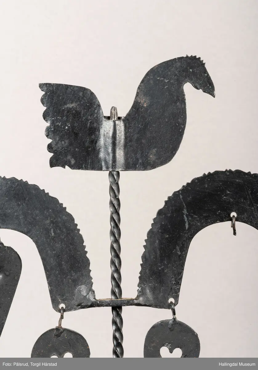 En hanestake i sort jern. Er hjerteformet med bærestang i midten, på toppen av stangen er det plasset en hanefigur. Øverste del av hjerteformen har ruglete kanter, under den ene siden henger et dråpeformet påheng, denne mangler på andre siden. Nærmere midtstangen henger en dråpeformet påheng med hjerteformet hull på hver side. To lyseholdere er montert på hver side av hjerteformen, begge har dråpeformet påheng under. Nedre del av hjerteformen har trappformet utforming der det er montert 7 dråpeformet påheng, 3 på ene siden og 4 på den andre. Midtstangen er litt bredere nederst og er montert på fire bredere stetter. 