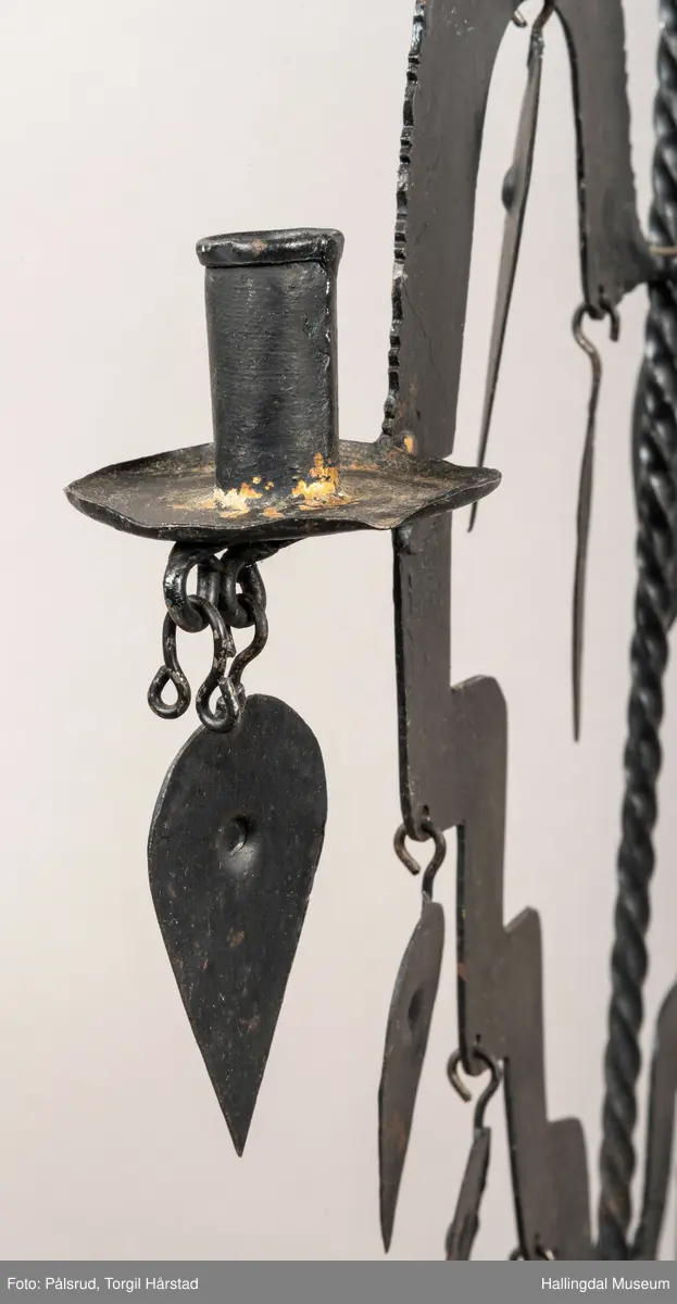 En hanestake i sort jern. Er hjerteformet med bærestang i midten, på toppen av stangen er det plasset en hanefigur. Øverste del av hjerteformen har ruglete kanter, under den ene siden henger et dråpeformet påheng, denne mangler på andre siden. Nærmere midtstangen henger en dråpeformet påheng med hjerteformet hull på hver side. To lyseholdere er montert på hver side av hjerteformen, begge har dråpeformet påheng under. Nedre del av hjerteformen har trappformet utforming der det er montert 7 dråpeformet påheng, 3 på ene siden og 4 på den andre. Midtstangen er litt bredere nederst og er montert på fire bredere stetter. 