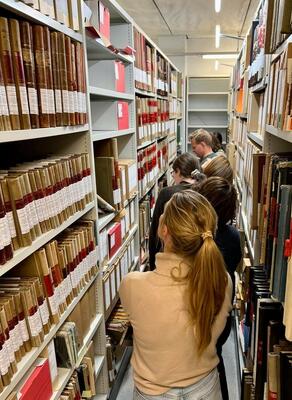 Rullearkiv, tre - fire personer som står mellom fulle arkivhyller på hver side av seg, og ser på innholdet. Nærmest kamera en kvinne med ryggen til, som ser innover i det smale rommet mellom de to hyllene.