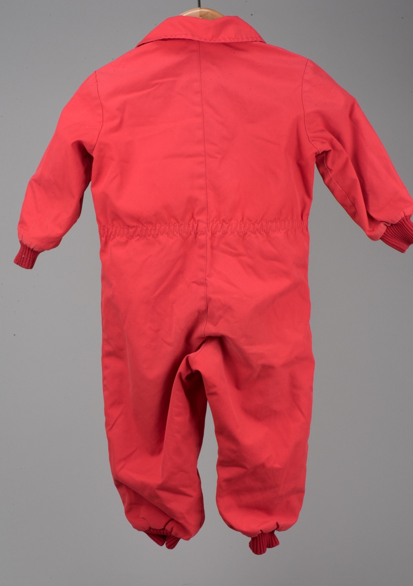 Röd bomullsoverall för barn i storlek 91.
Overallen har dubbla dragkedjor på framsidan, ärm- och benmuddar samt en rundad krage. I midjan finns ett insytt resårband.

På en etikett inuti overallen finns skötselråd men också Kooperativa förbundets symbol, oändlighetssymbolen en liggande åtta, samt "Babynett".

Overallen är välanvänd med slitna knän. De små cirkelformade märkena på overallen kommer från maskrosstjälkar. I grenen är overallen lagad, med handsydda stygn. Resåren i midjan har tappat sin spänst.