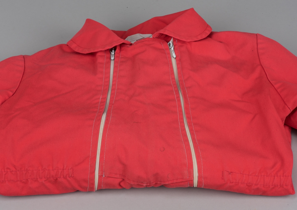 Röd bomullsoverall för barn i storlek 91.
Overallen har dubbla dragkedjor på framsidan, ärm- och benmuddar samt en rundad krage. I midjan finns ett insytt resårband.

På en etikett inuti overallen finns skötselråd men också Kooperativa förbundets symbol, oändlighetssymbolen en liggande åtta, samt "Babynett".

Overallen är välanvänd med slitna knän. De små cirkelformade märkena på overallen kommer från maskrosstjälkar. I grenen är overallen lagad, med handsydda stygn. Resåren i midjan har tappat sin spänst.