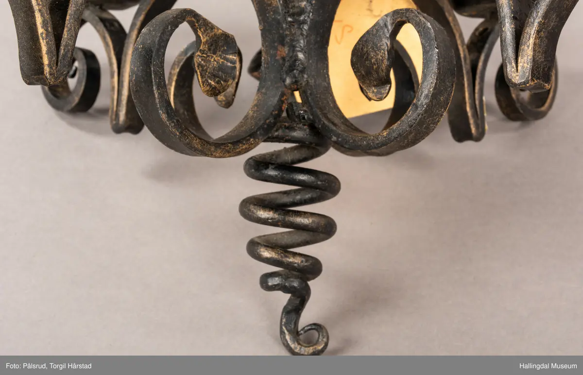 En firearmet lysekrone i sort smijern. Fire runde lysholdere. Metallet er sort med pusset gullfarge. En papirlapp er festet til lysekronen med hampetau. 