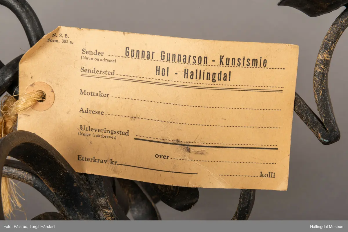 En firearmet lysekrone i sort smijern. Fire runde lysholdere. Metallet er sort med pusset gullfarge. En papirlapp er festet til lysekronen med hampetau. 