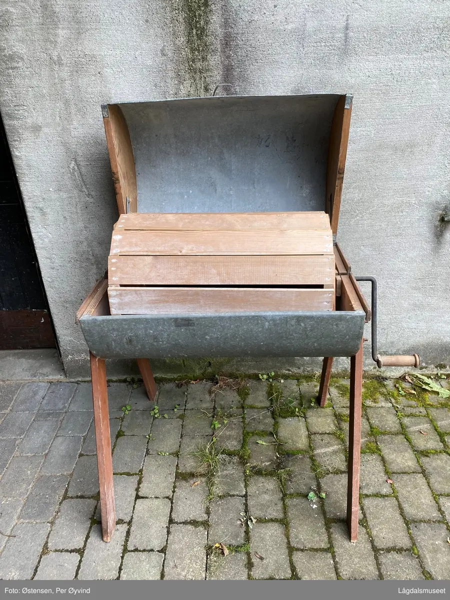 Hånddreven vaskemaskin utført i tre og blikk. Føtter, sider på vaskekassen og trommelen i kassen er utført i tre. Resten i blikk. Sveivestang i jern med dreid trehåndtak.  Vaskekassen er hengslet bak. Treverket er utvendig malt rødt og med logo for produsent på ene siden.