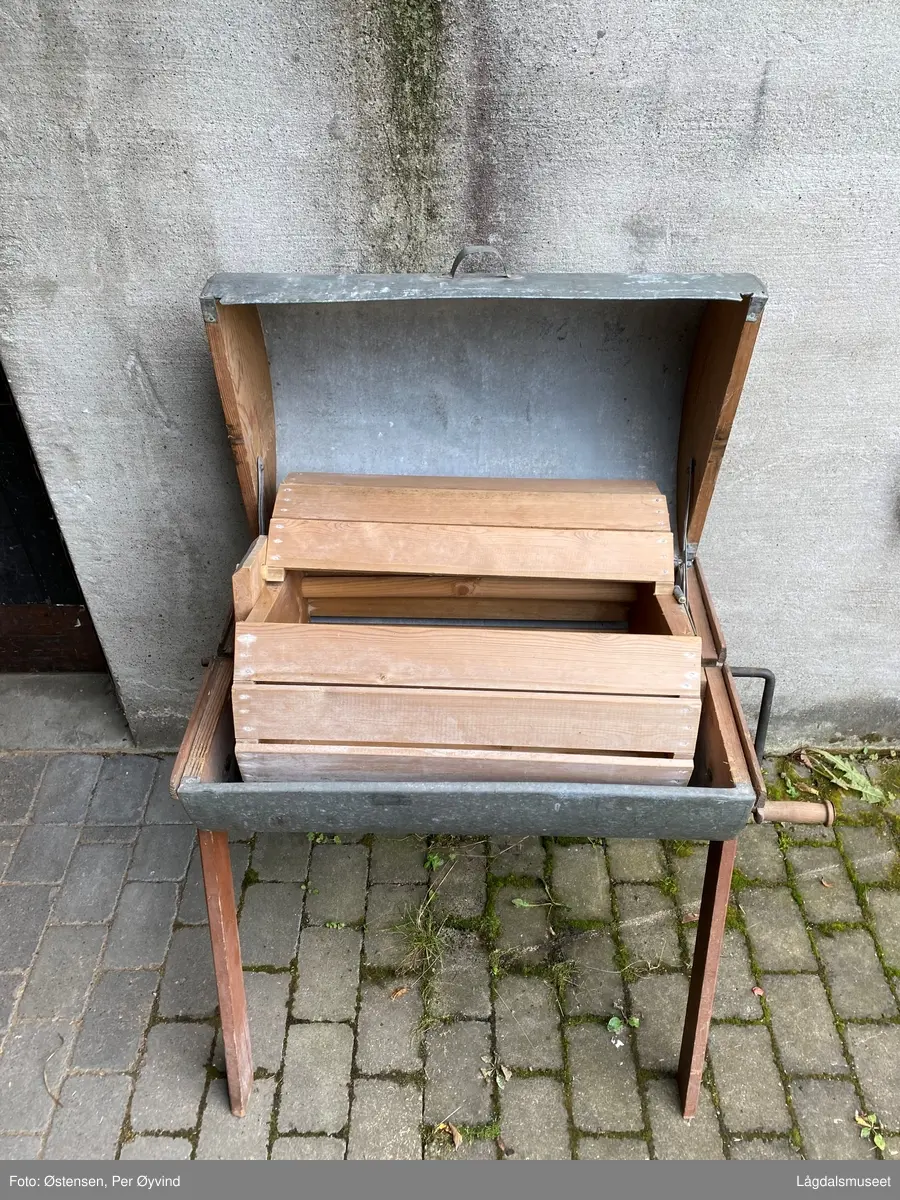 Hånddreven vaskemaskin utført i tre og blikk. Føtter, sider på vaskekassen og trommelen i kassen er utført i tre. Resten i blikk. Sveivestang i jern med dreid trehåndtak.  Vaskekassen er hengslet bak. Treverket er utvendig malt rødt og med logo for produsent på ene siden.