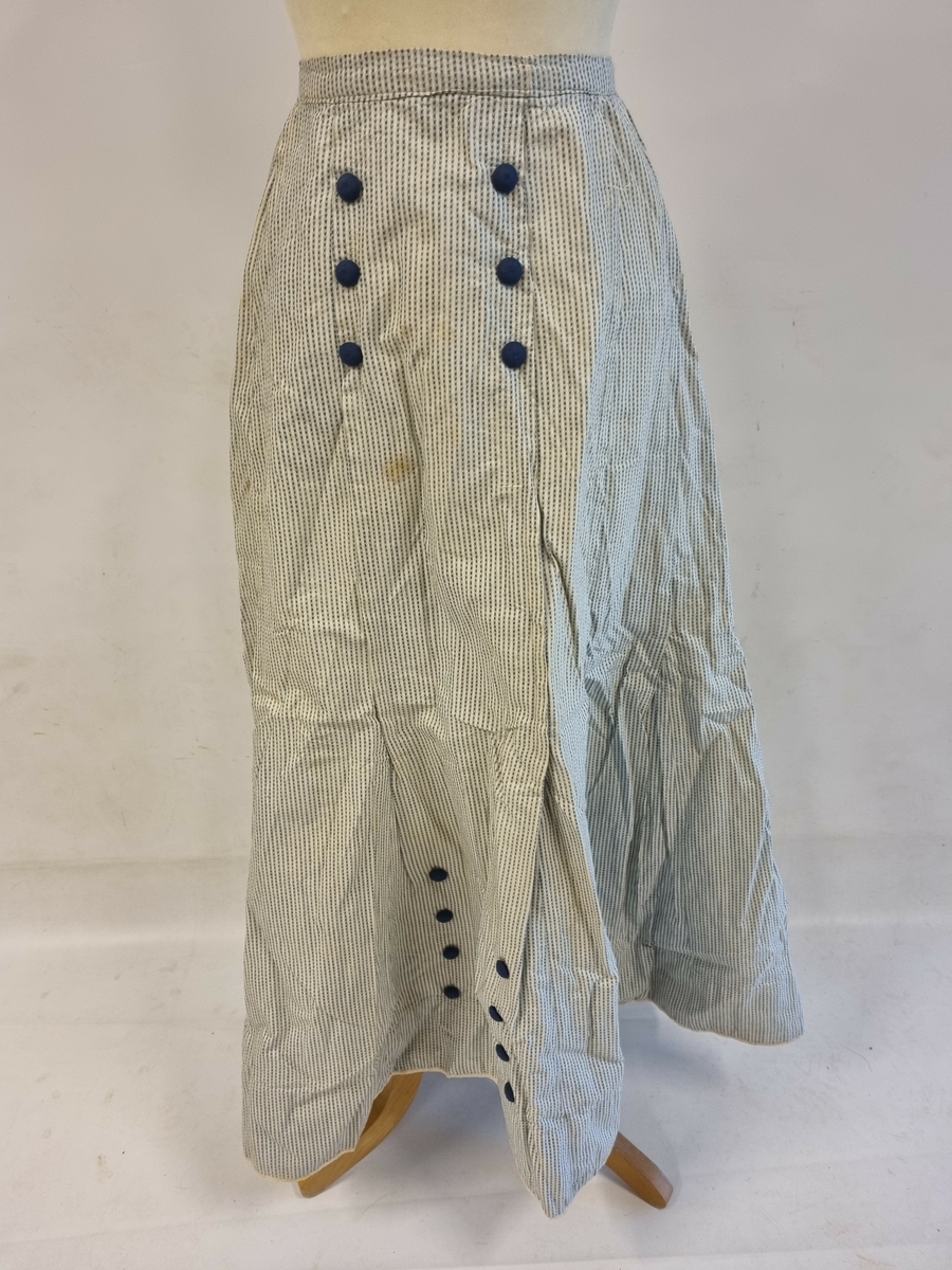 Langt skjørt med linning. Nedsydde folder. Pynteknapper: 6 stk. over magen samt 8 stk nederst. Knappene er stofftrukne. Lukning bak med hekter og trykknapper.