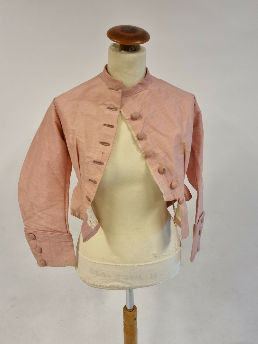 Tettsittende med skjørt. Ermer med oppbrett og 3 trukne knapper. Lukning i front med 7 trukne knapper. Halslinning. Fòret. 2 stk pynteknapper i ryggen.