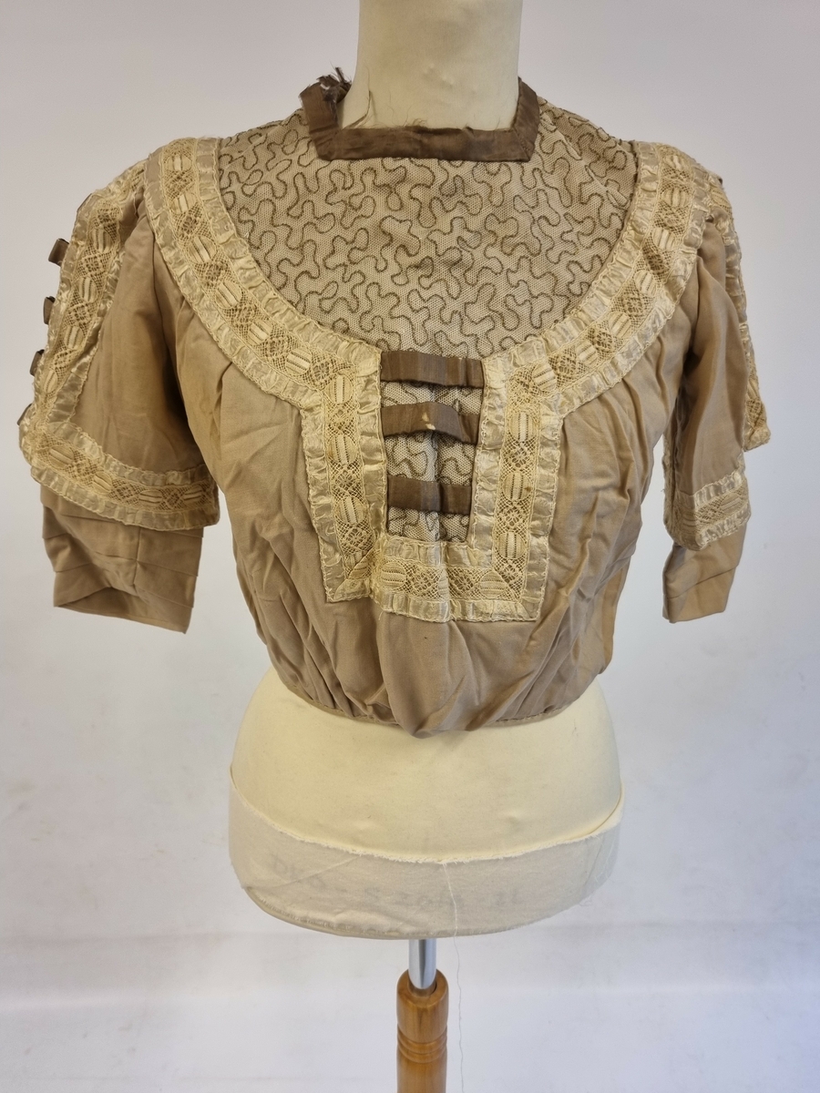 Bærestykke pyntet med blonder og silkebånd. Ermerlagt i leggmed pynt av blonder og silkebånd. Firskåren halsringing og lukking med trykknapper og hekter i ryggen. Spiler. Foret.