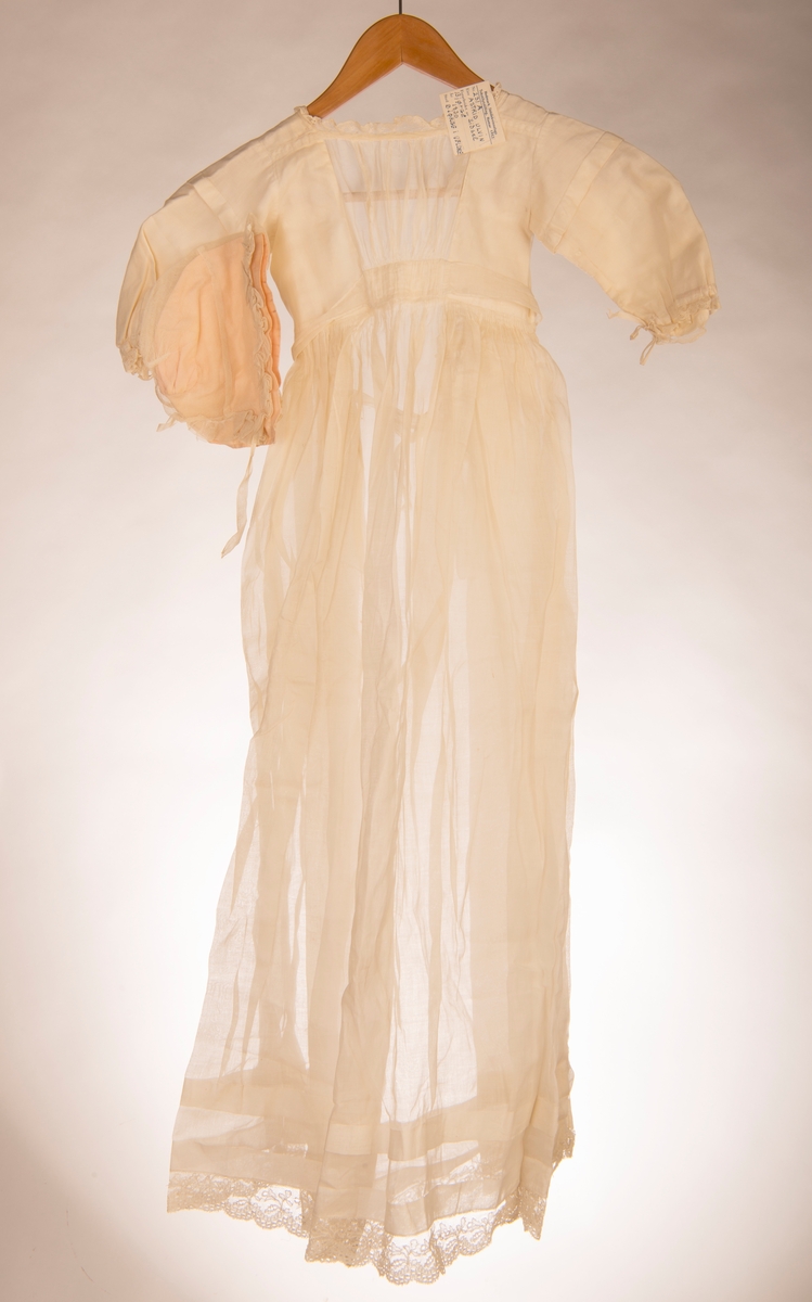 Dåpskjole med lange ermer. Fòret liv og påsydd rynket skjørt. Knytebelte i livet. Hals, ermer og skjørt kantet med kniplinger. Det er legg på skjørtet.
