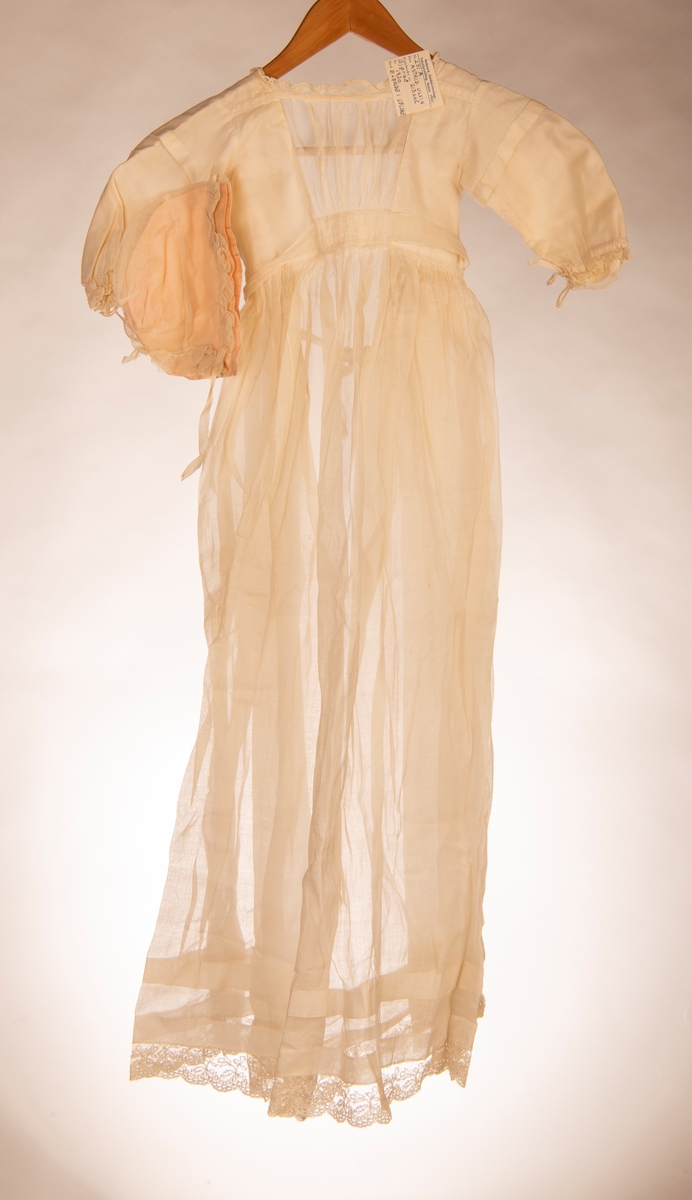 Dåpskjole med lange ermer. Fòret liv og påsydd rynket skjørt. Knytebelte i livet. Hals, ermer og skjørt kantet med kniplinger. Det er legg på skjørtet.
