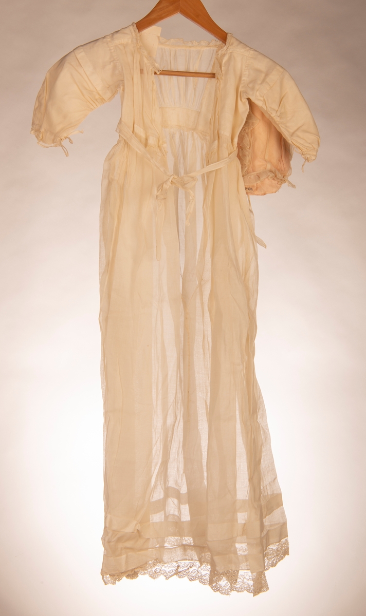 Dåpskjole med lange ermer. Fòret liv og påsydd rynket skjørt. Knytebelte i livet. Hals, ermer og skjørt kantet med kniplinger. Det er legg på skjørtet.
