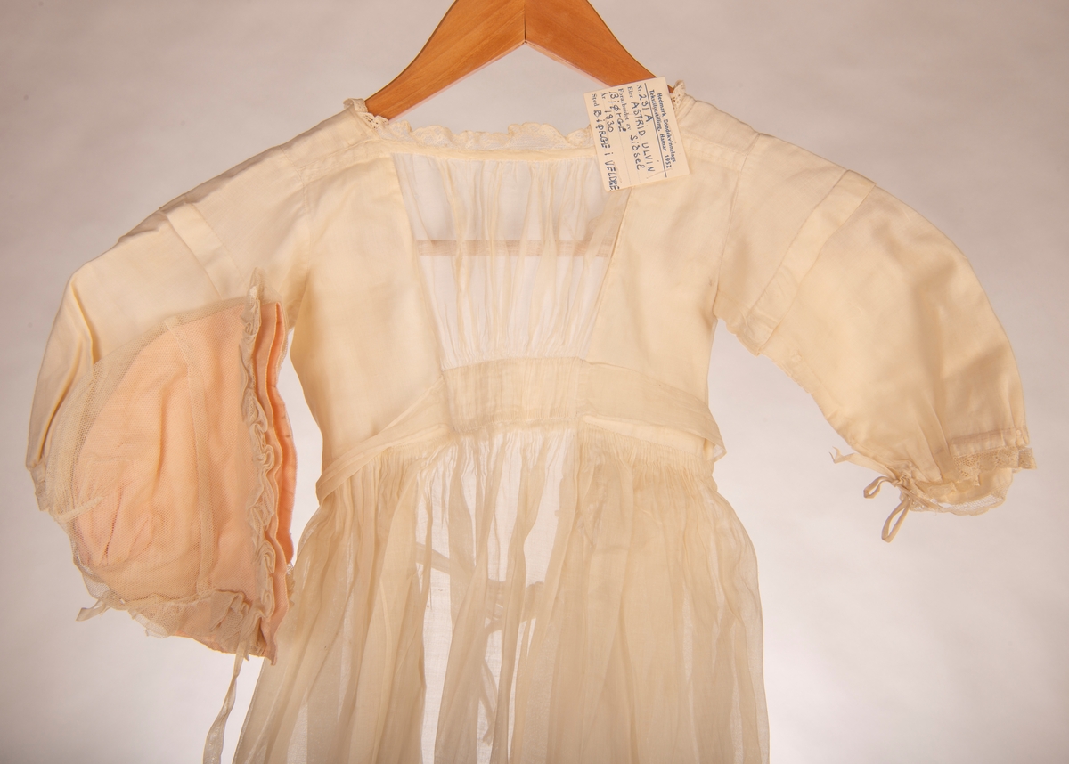 Dåpskjole med lange ermer. Fòret liv og påsydd rynket skjørt. Knytebelte i livet. Hals, ermer og skjørt kantet med kniplinger. Det er legg på skjørtet.
