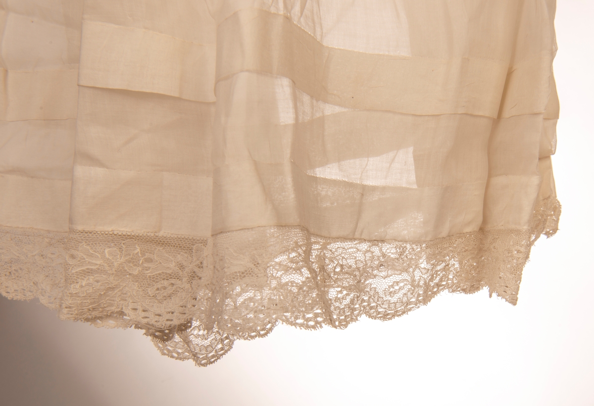 Dåpskjole med lange ermer. Fòret liv og påsydd rynket skjørt. Knytebelte i livet. Hals, ermer og skjørt kantet med kniplinger. Det er legg på skjørtet.
