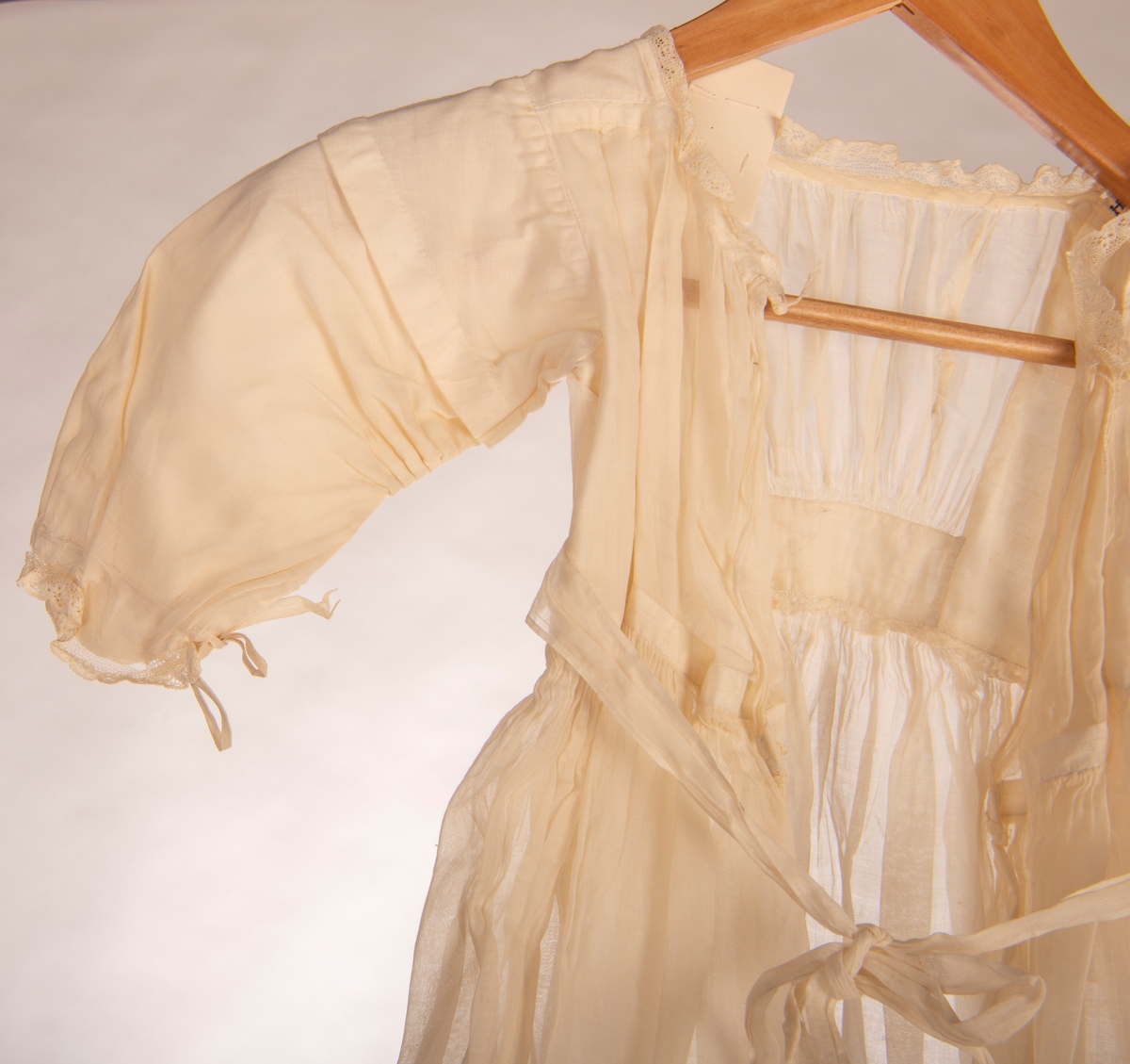 Dåpskjole med lange ermer. Fòret liv og påsydd rynket skjørt. Knytebelte i livet. Hals, ermer og skjørt kantet med kniplinger. Det er legg på skjørtet.
