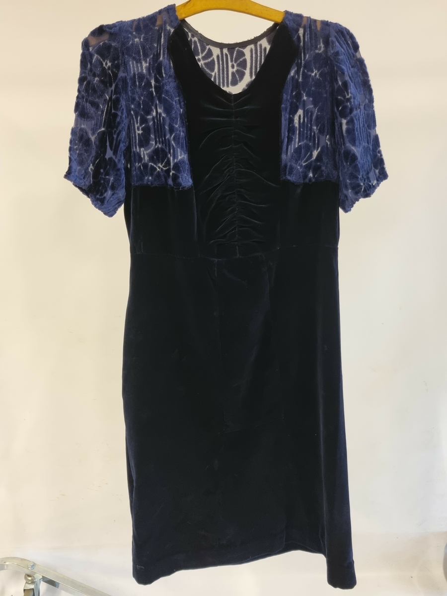 Selskapskjole med båtformet halsringning og korte ermer. Bærestykke i mønstervevet fløyel kan være del av omsøm, resten er i mørkeblå bomullsfløyel. Hjemmesydd eller fra sydame. Fra ca. 1930-tallet.