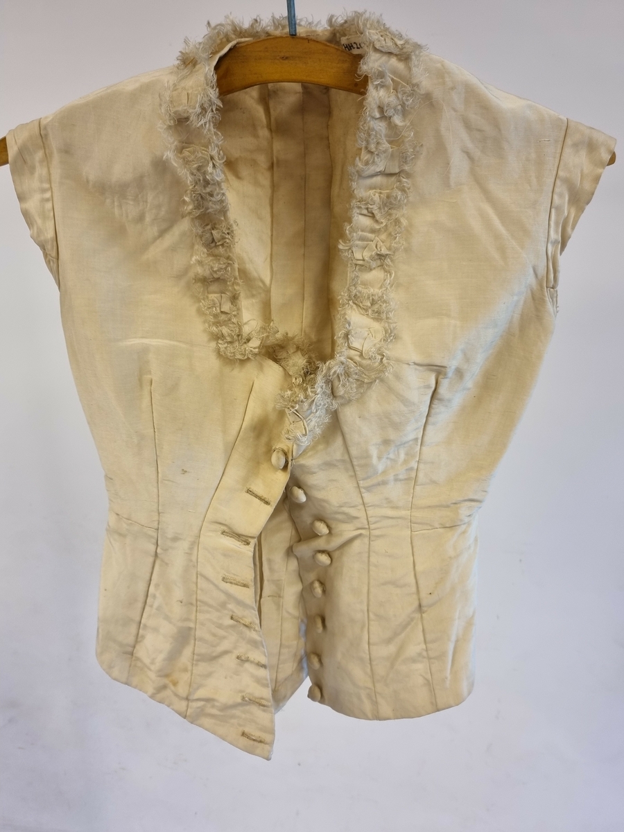 Hvitt kjoleliv i bomull, trukket med organza. Ermeløst, og med en utringning pyntet med små sløyfer og frynser. Forknappet med trukne knapper. Skjult belte.