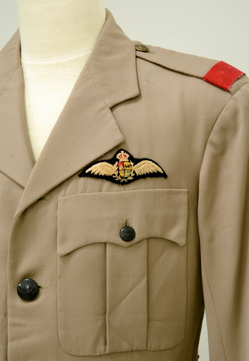 A.Uniformsjakke. Tropeuniform. Beige jakke i syntetisk materiale med 4 lommer foran. 8 knapper med emblem, skulderklaffene med rødt merke uten distinksjoner. Påsydd flygersymbolmerke foran. Jakken har fôr i armene og ved nakkepartiet. Belte med beltespenne.

B. Uniformslue. Luen har en brett festet med trykk-knapp på begge sider. Denne bretten kan brettes ut, og det blir en lue som kan festes under haken med to små knapper med emblem. Innvendig er det svettebånd og fôr.