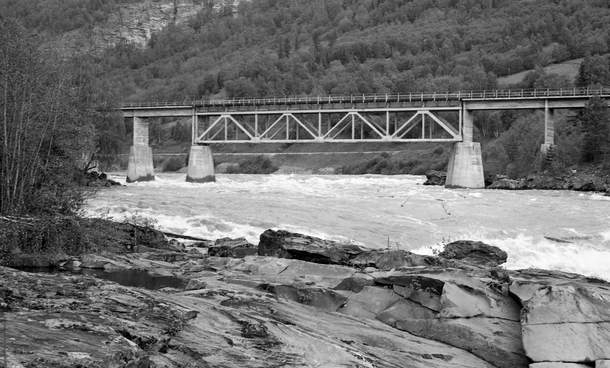 Brua over den øvre delen av Solbråfossen i Gudbrandsdalslågen, fotografert i motstrøms retning fra ei bergflate på vestre elvebredd. Denne lokaliteten ligger i Sør-Fron kommune i Gudbrandsdalen. Brua ble antakelig bygd i forbindelse med reguleringa av fossen, og den var følgelig ganske ny da dette fotografiet ble tatt i 1963. Det dreier seg om ei plate- eller bjelkebru, som i hovedsak er utført i armert betong, men der det største spennet, som er plassert over djupålen i elva, er en fagverkskonstrukasjon med stålelementer. I bakgrunnen ser vi blandingsskogen oppover i Skarslia.