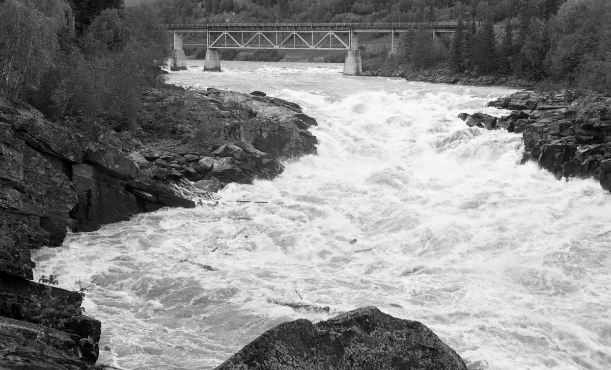 Brua over den øvre delen av Solbråfossen i Gudbrandsdalslågen, fotografert i motstrøms retning fra vestre elvebredd et par hundre meter nedenfor. Denne lokaliteten ligger i Sør-Fron kommune i Gudbrandsdalen. Brua ble antakelig bygd i forbindelse med reguleringa av fossen, og den var følgelig ganske ny da dette fotografiet ble tatt i 1963. Det dreier seg om ei plate- eller bjelkebru, som i hovedsak er utført i armert betong, men der det største spennet, som er plassert over djupålen i elva, er en fagverkskonstrukasjon med stålelementer. I bakgrunnen ser vi blandingsskogen oppover i Skarslia.