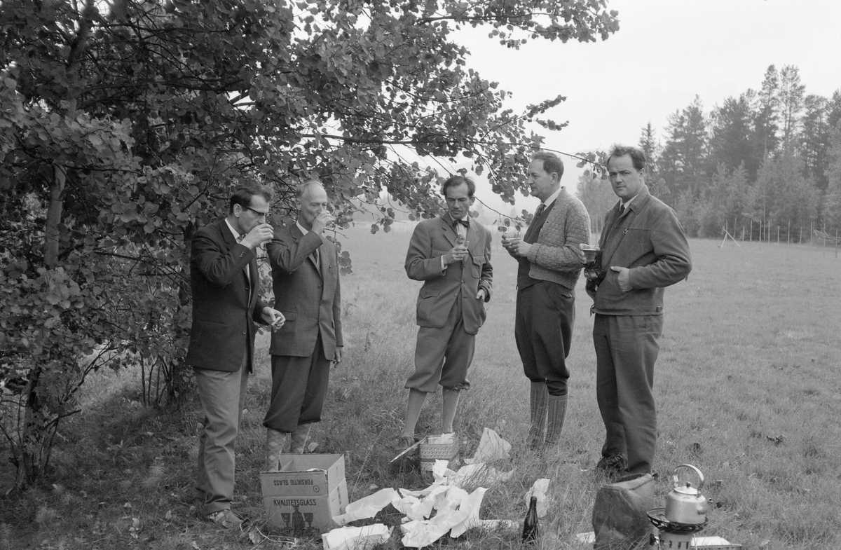 Fem fløtingsfunksjonærer, fotografert under befaring, antakelig ved Trysilvassdraget. Fotografiet er tatt i utkanten av ei eng, under noen lauvtrær i randsona, der mennene inntok et lunsjmåltid stående. Fotografen har notert seg navnene Modin, Sørebø, Svensson, Akre og Svensson. Mannen lengst til høyre er gjenkjent som Per Ivar Akre (1925-1997). Han var andre generasjons fløtersjef i den norske delen av vassdraget, noe faren Paul Akre (1888-1952) også hadde vært.