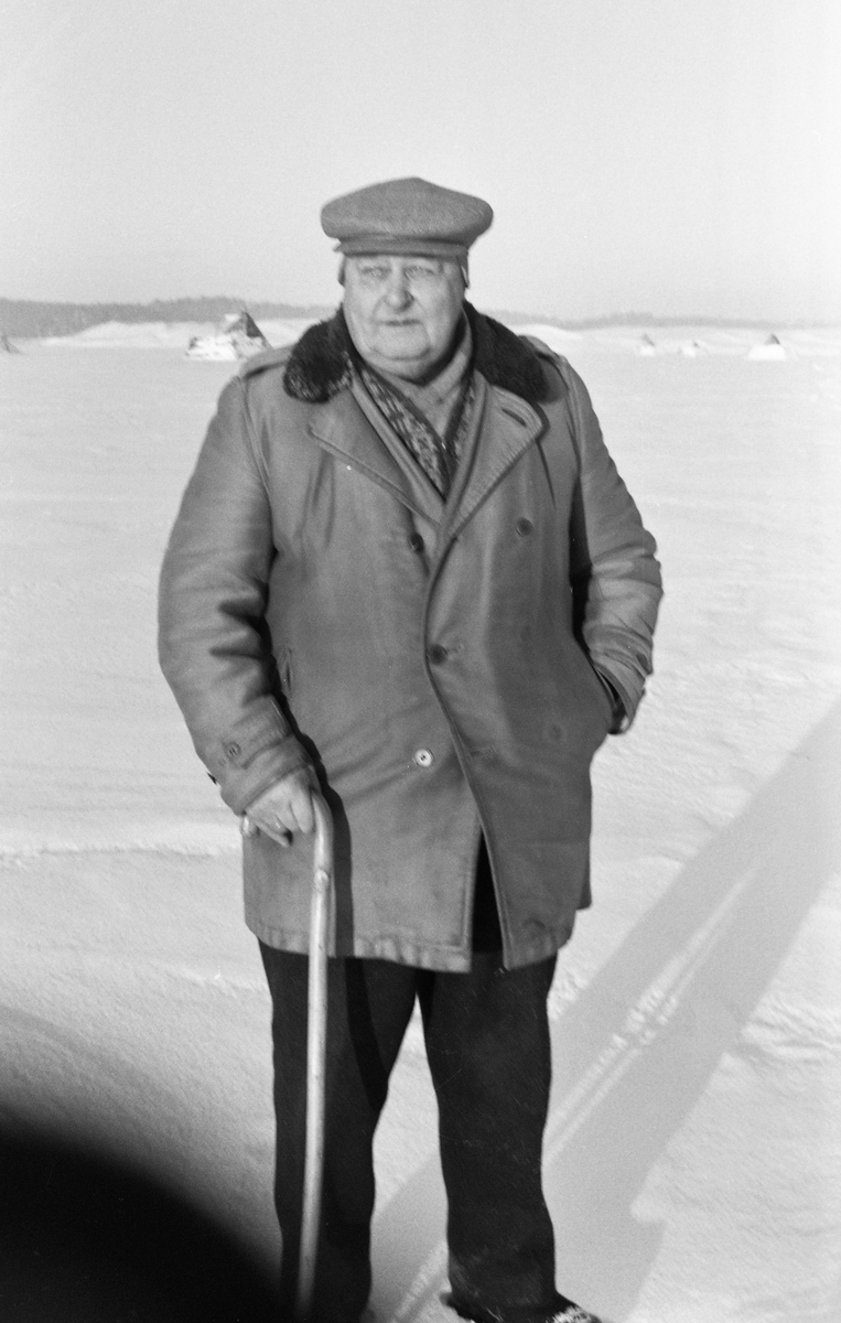 Fløterveteranen Adolf Vestby (1894-1975), mangeårig formann ved Glomma fellesfløtingsforenings attholdslense ved Bingen i Sørum, Fotografiet er tatt vinteren 1963 på det is- og snødekte elveløpet som var Vestbys arbeidsplass i sommersesongen. Fløterformannen var kledd i ei tjukk vinterjakke og hadde sixpencelue med ørelapper på hodet. I den ene handa holdt han en spaserstokk, den andre varmet han i jakkelomma. I bakgrunnen skimter vi de såkalte riarkarene ved lenseanlegget.