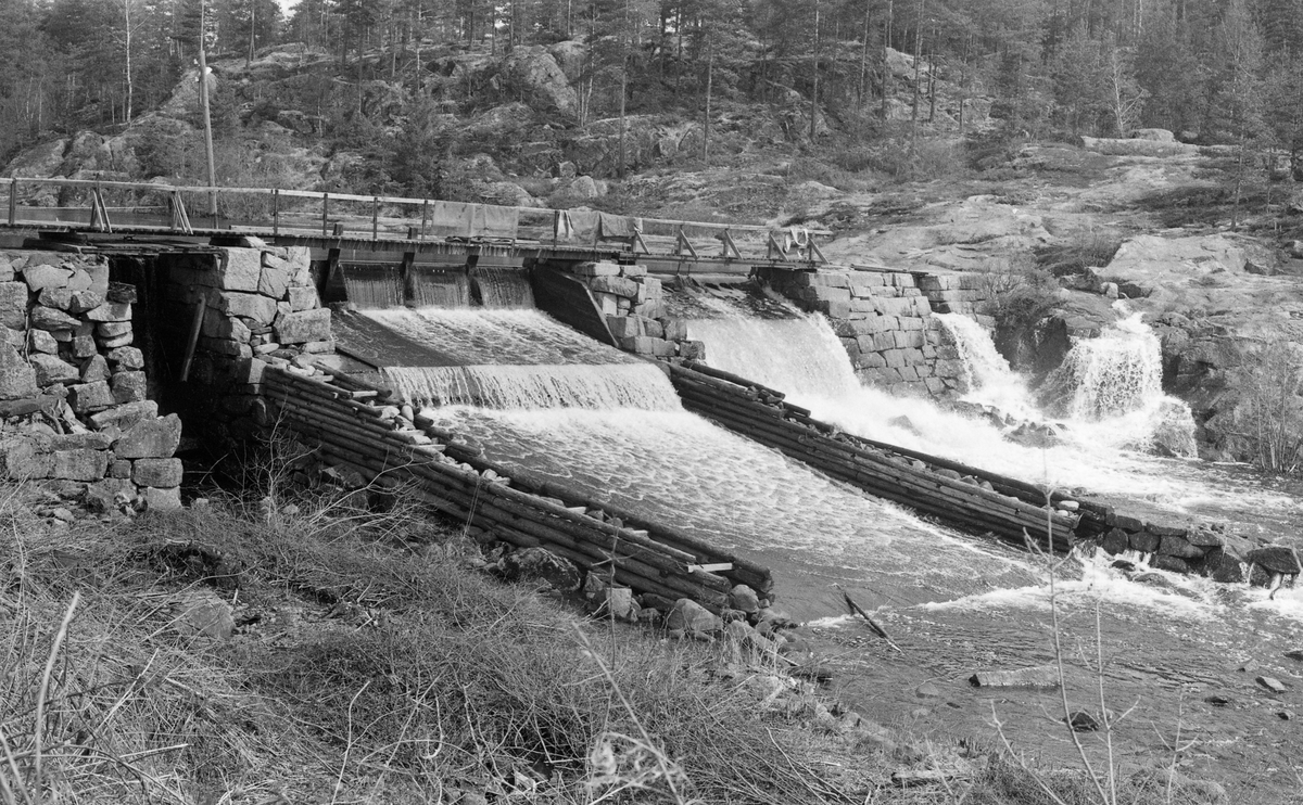 Klevfosdammen i Svartelva, på Ådalsbruk i Løten, Hedmark. Fotografiet er tatt fra vest mot øst, skrått på strømretningen. Denne dammen var et vannreservoar for Klevfos Cellulose- & Papirfabrik (1888-1976), der maskinene dels ble drevet av vannkraft, dels av en diger dampmaskin. Dammen er murt av kvadret naturstein. Den har to forholdsvis brede løp i den sentrale delen av elveløpet og to smalere åpninger i ytterendene. Det smale løpet helt til venstre ble antakelig brukt når det var behov for å tømme dammen slik at det ble mulig å gjøre repearasjonsarbeider. Det brede løpet ved siden av denne var utstyrt med steinfylte tømmerkasser som avgrensninger mot sidene. Over det hele går det ei trebru mellom søndre og nordre elvebredd. Dette fotografiet ble tatt i 1964,